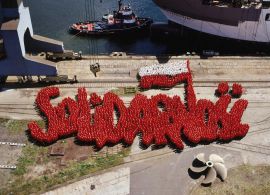 1	część dyptyku. Zdjęcie przedstawia kilka tysięcy żołnierzy, ubranych  w czerwone i białe bluzy, ustawionych w taki sposób, że tworzą znak Solidarności. Dominuje czerwień, biel pojawia się w polskiej fladze. Stoją na betonowym nabrzeżu.