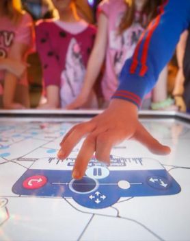Dzieci w  pomieszczeniu przy interaktywnej mapie. Jedno z dzieci dotyka mapy dłonią.