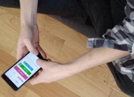 Fragment postaci widzianej z góry. Siedzi po turecku. Czarne spodnie, koszula w kratę. Dłonie trzymają smartfona. W tle drewniana podłoga.