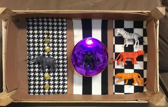 Na zdjęciu widzimy drewniane pudełko wyłożone od wewnątrz 3 kawałkami  materiałów, w różnego rodzaju biało-czarne wzory, a na nich kolejno od lewej - czarna figurka słonnia i 5 złotych guzików, okrągła foremka wyłożona fioletowym papierem, a na środku gor