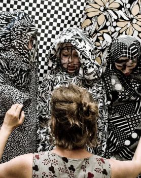 3 dzieci, z pomalowanymi twarzami, okryta materiałami w kamuflażowym nadruku oraz kobieta, która kuca przed nimi (widzimy ją tyłem). W tle wiszą materiały w czarno-białą kratę i kwiaty.