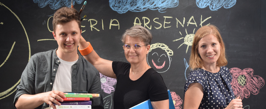 Na zdjęciu widoczni są uśmiechnięci pracownicy działu edukacji Galerii Arsenał. Od lewej - Aleksander trzymający książki, Kasia trzymająca namalowany obraz przedstawiający łąkę, oraz Justyna mająca bransoletki z rolek kolorowych taśm klejących. W tle nary