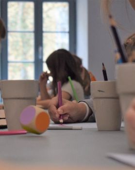 Sala warsztatowa. Dzieci rysują kredkami. Po stole toczy się kolorowa kostka.