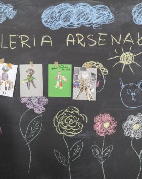 Na czarnej ścianie napis kredą GALERIA ARSENAŁ. Wokół rysunki i prace dzieci. Po lewej dziewczynka wiesza swoją pracę.