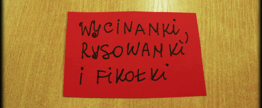 na drewnianym blacie leży czerwona kartka, na której napisano czarnym mazakiem "wycinanki, rysowanki i fikołki"