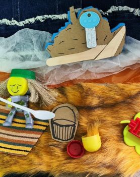 Praca plastyczna - asamblaż pokazuje chłopca na kocu na plaży nad brzegiem morza. Praca stworzona z różnych materiałów i przedmiotów np. futro, korek, łyżeczka, stara zabawka, karton.
