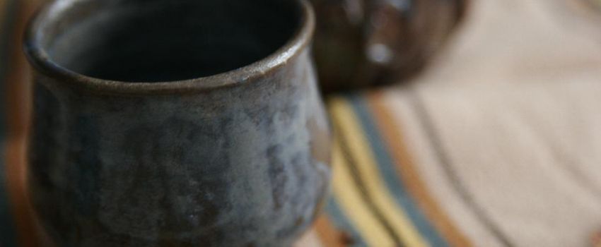 Na kolorowej tkaninie stoi gliniany kubek w kolorze brązowo- niebieskim. W tle inne naczynie gliniane.