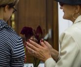 Dwie osoby stojące tyłem. Widać jedynie fragment ramienia i głowy każdej z nich. Obie noszą okulary. Osoba po lewej stronie ma niebieską bluzkę w granatowe paski. Osoba po prawej ma beżową marynarkę, nosi kapelusz. Jest uśmiechnięta. Ma ręce zgięte w łokc