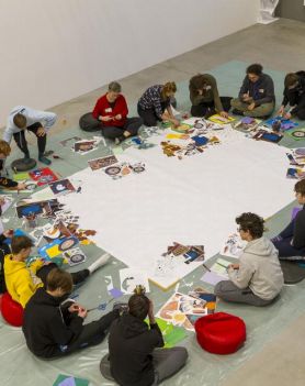 Widok z góry z 3/4. Grupa młodzieży siedzi na podłodze w okręgu wokół dużego białego papieru. Wycinają wydrukowane elementy i przyklejają na kolorowe kartki.