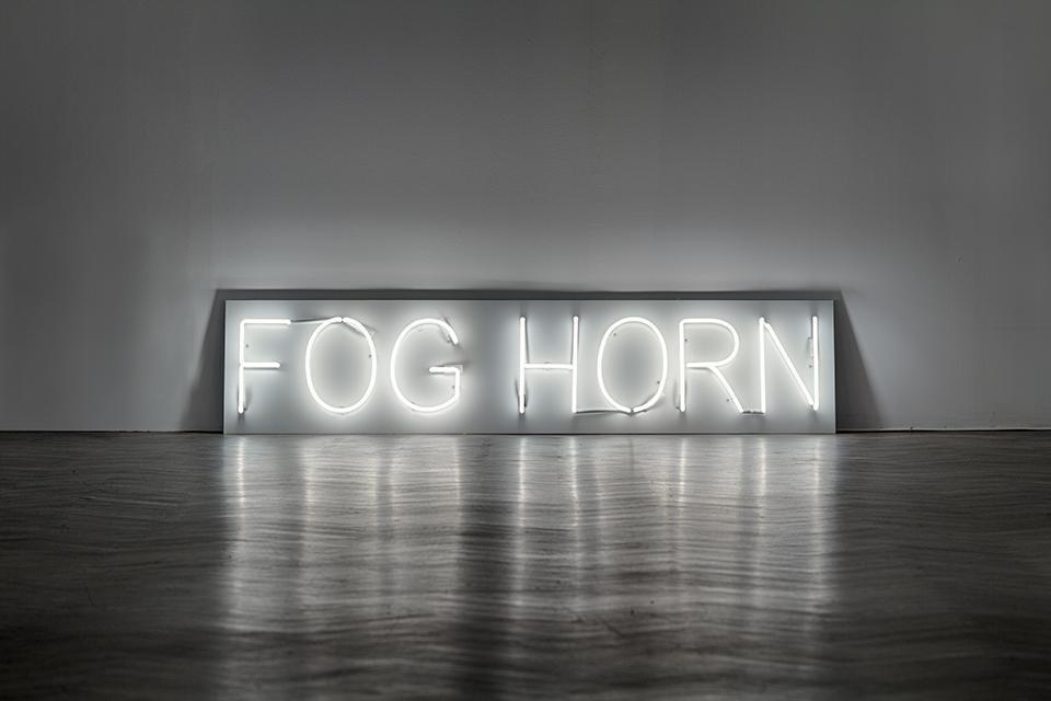 Neon stojący na drewnianej podłodze i oparty o białą ścianę. Świeci zimnym jasnym światłem. Przedstawia napisy wielkimi literami na jasnym prostokącie.