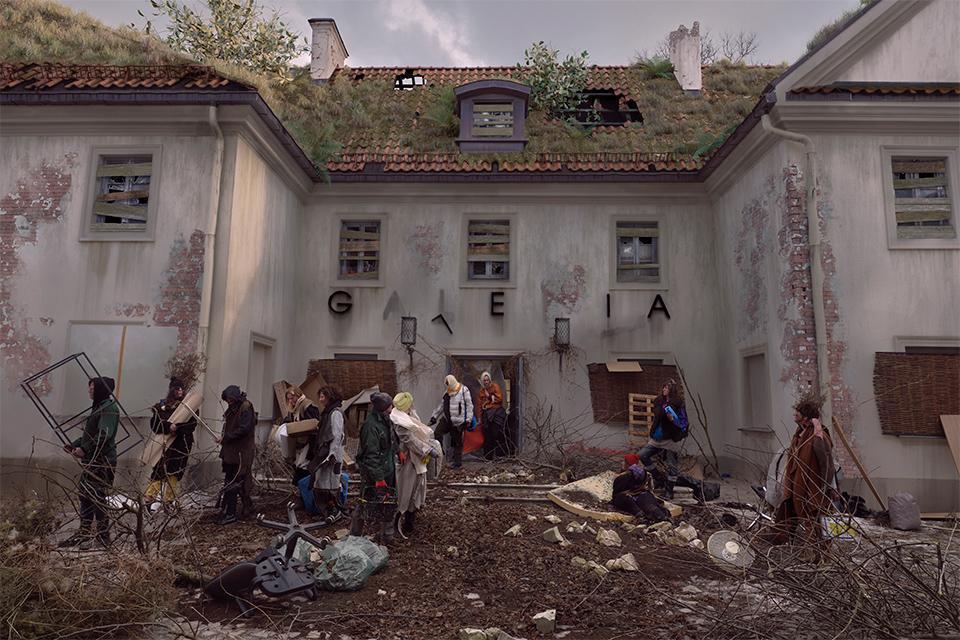 Zniszczony budynek z dachem porośniętym mchem i roślinami. Przed nim grupa ludzi w zniszczonych ubraniach. Wokół panuje bałagan.