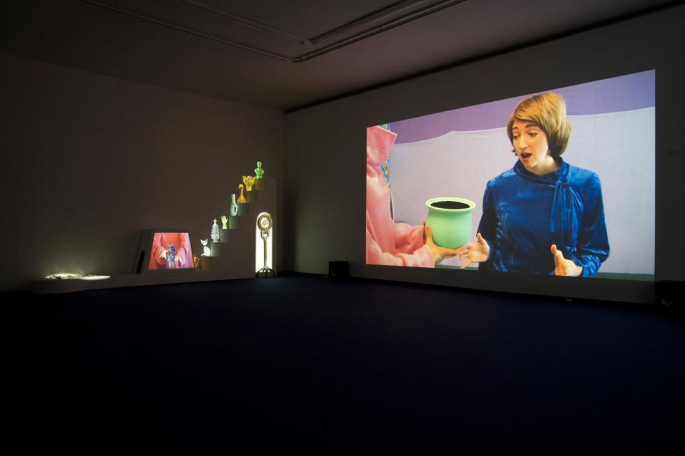 Róg nieoświetlonego pomieszczenia. Po prawej stronie projekcja wideo wyświetlana na ścianie. Po lewej kilka świecących przedmiotów.