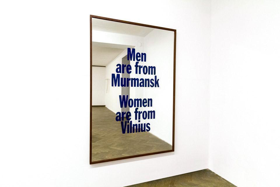 Lustro w ramie z ciemnego drewna. Na powierzchni szkła widnieje sześć wersów ciemnoniebieskiego tekstu. Lustro wisi na białej ścianie. Odbija się w nim reszta pomieszczenia z drewnianą podłogą.