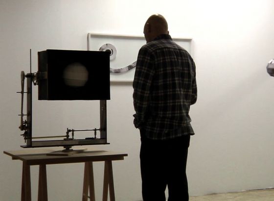 Mężczyzna stoi przed instalacją usatwioną na drewnianym stoliku, na tle białej ściany na której widać fragmenty obrazu. Stoi on tyłem do obiektywu aparatu.