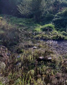 Instalacja znajdująca się na wolnym powietrzu. Ustawiona jest na terenie piaszczystym, porośniętym roślinami i trawami.