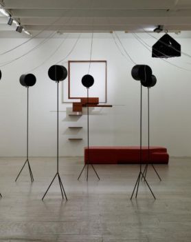 W pomieszczeniu o białych ścianach, na środku widoczna jest instalacja. Druga jej część znajduje się na ścianie. Przy ścianie ustawione jest prostokątne siedzisko w kolorze czerwonym.