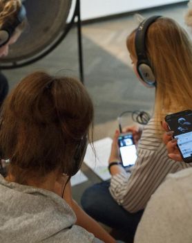 Cztery kobiety trzymające telefony komórkowe. Na uszach mają słuchawki. siedzą na podłodze wpomieszczeniu. Zdjęcie zrobione z góry.