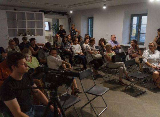 W sali, na krzesłach siedzą ludzie. Dwie kobiety stoją na końcu rzędu, tuż przy wejściu do pomieszczenia. W tle biały regał z szafkami i półkami.