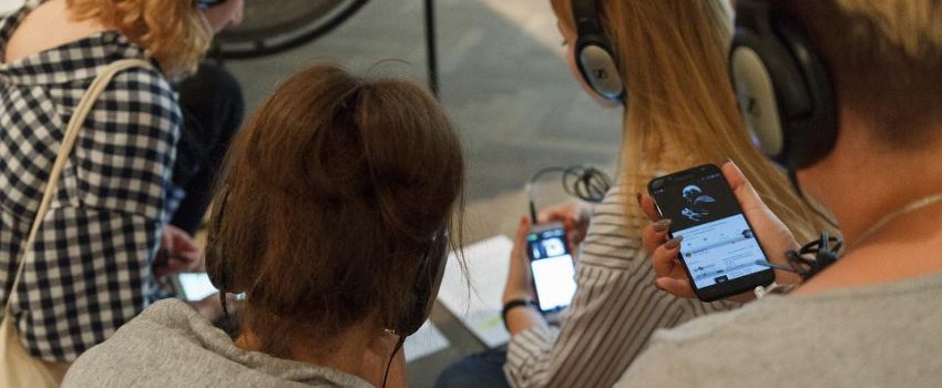 Młodzież siedzi na podłodze podczas warsztatów. W dłoniach trzymają telefony komórkowe, na uszach mają słuchawki. Zdjęcie zrobione z góry.