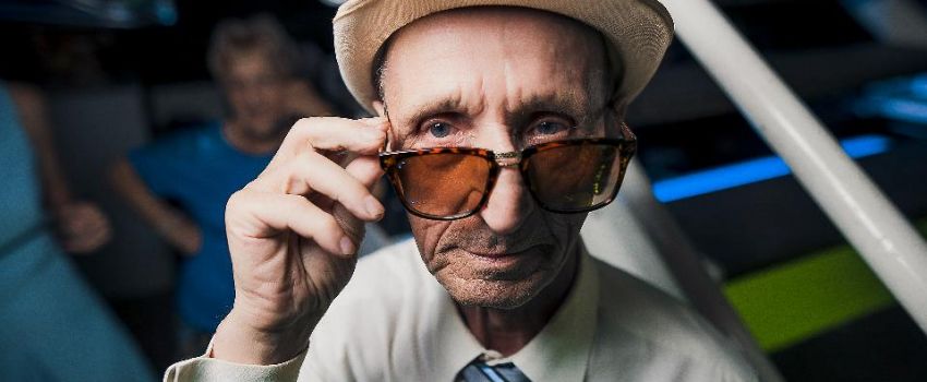 Na zdjęciu starszy mężczyzna w kapeluszu i okularach. Jedną dłonią dotyka oprawki okularów.