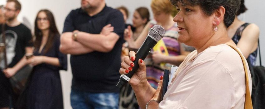 Na pierwszym planie widoczna kobieta, która przemawia do mikrofonu. W tle tłum ludzi.