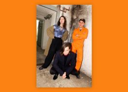 Na zdjęciu widoczna trójka ludzi. Dwie osoby stoją przy ścianie, jedna kuca. Zdjęciu umieszczone w pomarańczowej ramce.