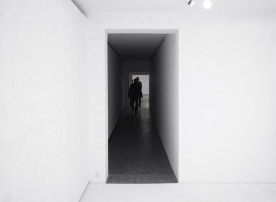 •	Białe pomieszczenie, od którego wychodzi wąski, zaciemniony korytarz prowadzący do innego pokoju. Widoczni przemieszczający się ludzie zwróceni tyłem do obiektywu aparatu.