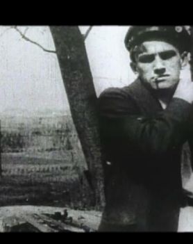 Czarno-biała fotografia. Na pierwszym planie mężczyzna, ukazany od pasa w górę. Ubrany w marynarkę, na głowie ma czapkę. W ustach trzyma papierosa.