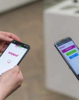 w centralnej części zdjęcia trzymane przez dwie różne osoby dwa telefony, na których widoczne jest logo gry SEPPO