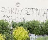na beżowej ścianie napis czarnym sprayem - znak anarchii, pod spodem: Czarny Sztandar 1902 - C.D.N. , na pierwszym planie zielone krzewy