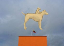 kadr z wideo na którym widzimy wygenerowaną komputerowo sylwetkę psa, któremu z grzbietu wyrasta popiersie mniejszego psa, w tle widać zachmurzone niebo i pomarańczowy budynek z masztem na którym powiewa flaga litewska