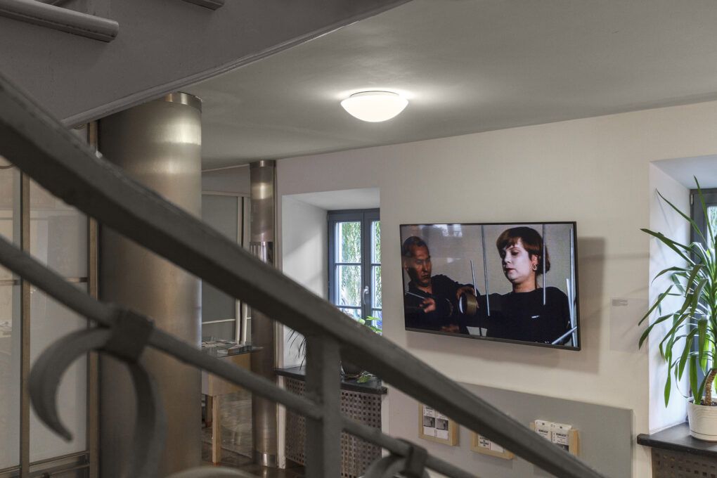 Projekcja wideo wyświetlana na telewizorze zawieszonym na białej ścianie. Po lewej stronie znajduje się okno. Po prawej widać część parapetu. Na pierwszym planie fragment balustrady.