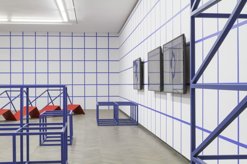Widok instalacji w przestrzeni Galerii Arsenał. Ściany pomieszczenia białe. Na nich namalowana niebieska kratka. Drewniana podłoga.