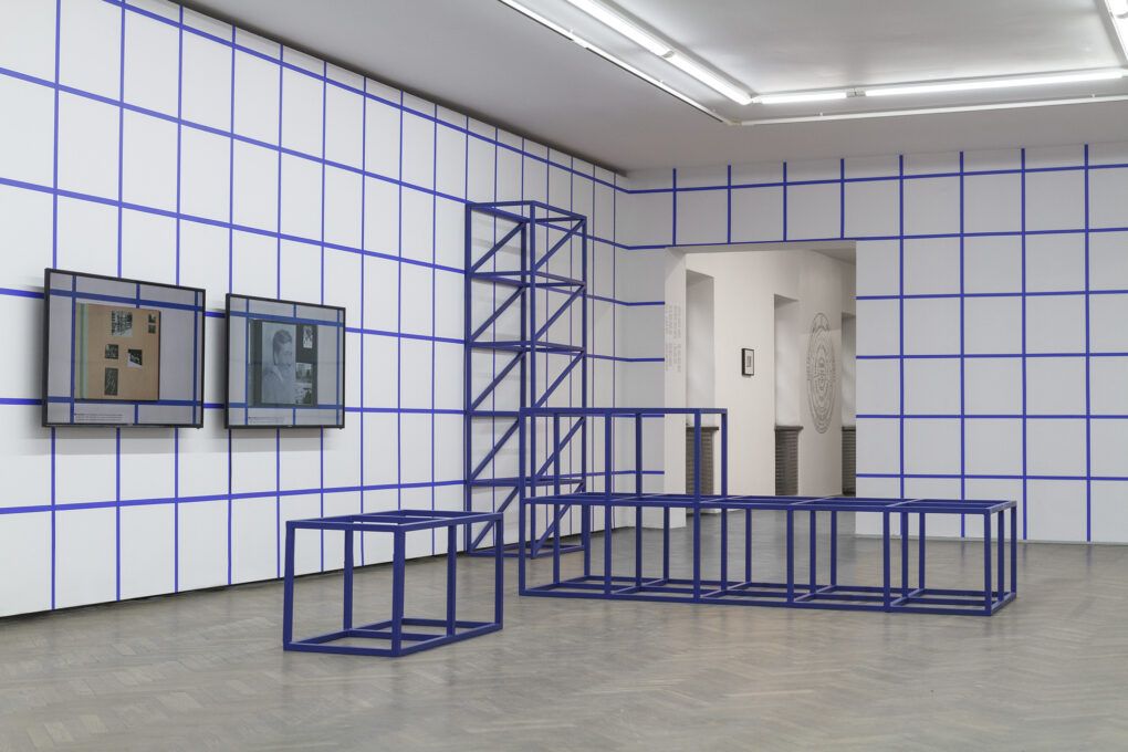 Widok instalacji w przestrzeni Galerii Arsenał. Ściany pomieszczenia białe. Na nich namalowana niebieska kratka. W tle wejście do kolejnej sali wystawowej.