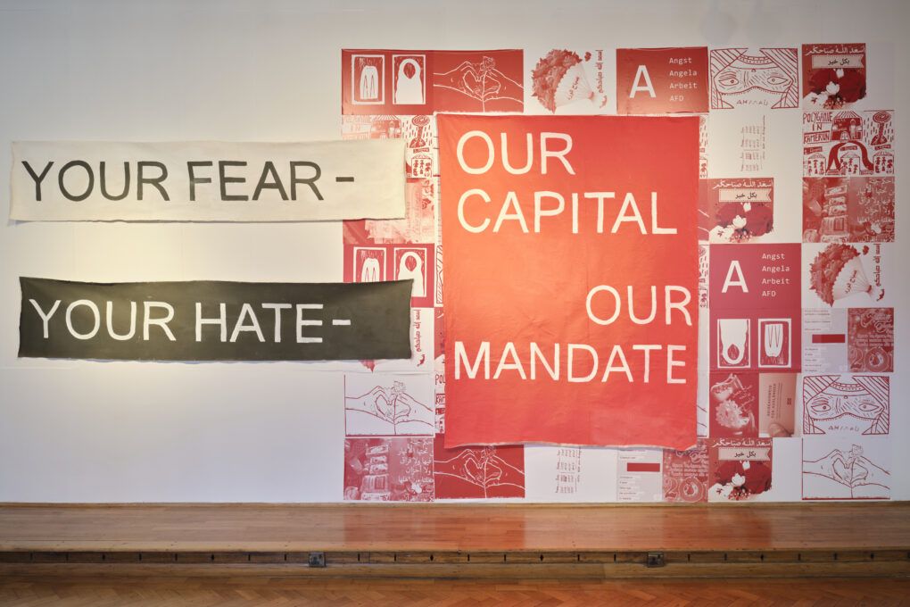 praca arystyna składająca się z wielkoformatowych kawałków materiału z napisami "your fear - our capital your hate - our mandate" oraz drugiej większej części gdzie w tle są białe i czerwone zdjęcia i napis